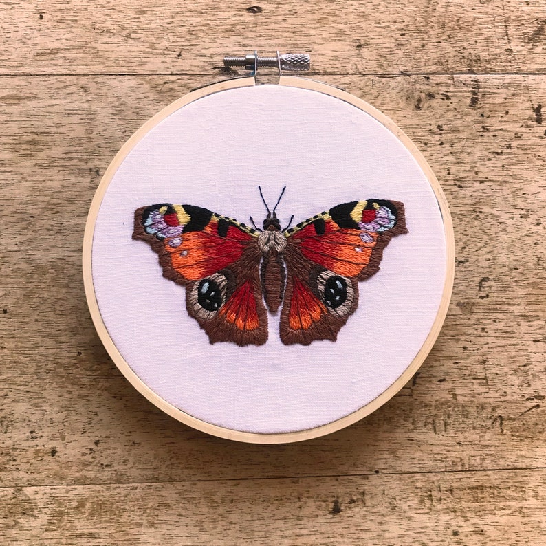 May include: Embroidered peacock butterfly on a white fabric background in a wooden embroidery hoop.