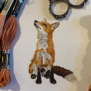 May include: A detailed embroidery of a red fox sitting up with its head tilted up. The fox is stitched with brown, white, and orange thread on a white fabric.