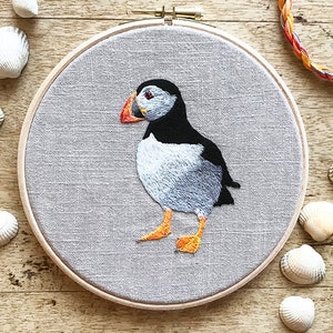 以下が含まれることがあります： 木製の刺繍枠の中に、グレーのリネン生地に刺繍されたパフィンが描かれています。パフィンは左を向いており、黒い頭、白い体、オレンジ色のくちばしと足を持っています。