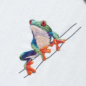 May include: An embroidered frog with red eyes, green head, and blue and purple body, perched on a black line against a white background. The frog's feet and toes are orange. The embroidery is detailed and colorful.