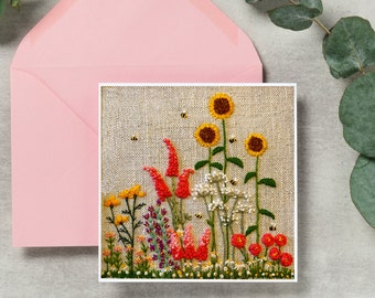 May include: A hand-stitched embroidery card featuring a colorful floral arrangement with red, orange, yellow, and white flowers. The flowers are surrounded by green leaves and bees. The card is on a light pink envelope.