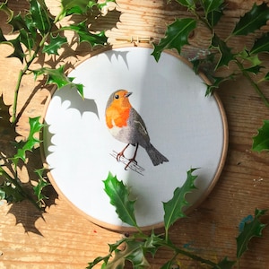 May include: Embroidered artwork of a robin perched on a branch, with a red breast and gray wings, set against a white background in a wooden embroidery hoop.