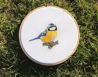 May include: A bluebird with a yellow breast and a blue head is embroidered on a white fabric stretched in a wooden embroidery hoop.