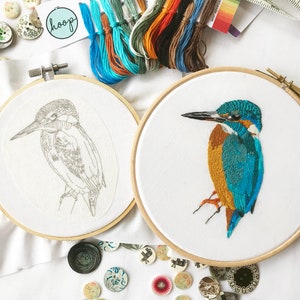 Puede incluir: Dos bastidores de bordado, uno con un dibujo en blanco y negro de un martín pescador y el otro con un martín pescador bordado en color. Los bastidores de bordado están sobre una superficie blanca con botones y otros materiales de artesanía.
