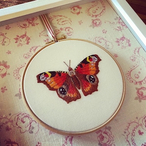 May include: A close-up of a framed embroidery of a peacock butterfly with brown, orange, and black wings. The embroidery is on a white background and is framed in a white frame.