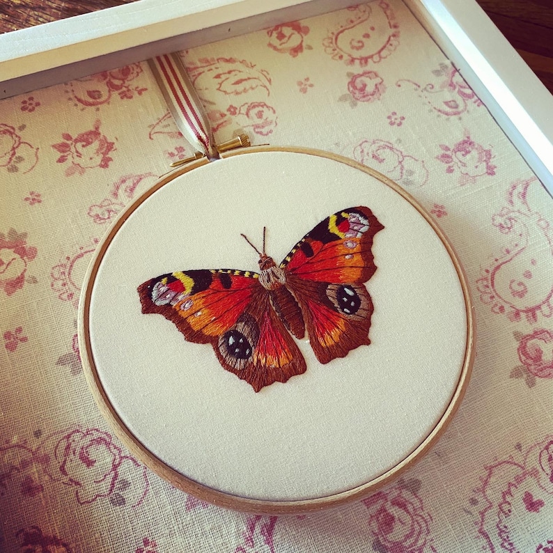 May include: A close-up of a framed embroidery of a peacock butterfly with brown, orange, and black wings. The embroidery is on a white background and is framed in a white frame.