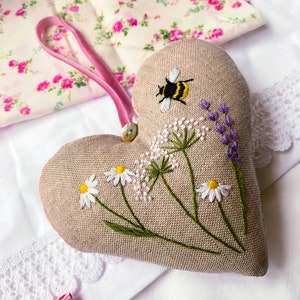 Könnte beinhalten: Ein herzförmiges Kissen aus Leinen mit gestickten Wildblumen, einer Biene und Lavendel. Das Kissen ist beige mit grünen Stielen und weißen und gelben Blüten.