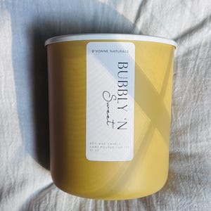 Op de afbeelding: Een gele sojawas kaars met een witte rand en een label met de tekst "Bubbly 'N Sweet". De kaars is met de hand gegoten en weegt 368.5 gram. De merknaam "D'Vonne Naturals" is ook zichtbaar.