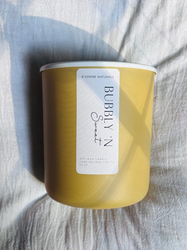 Op de afbeelding: Een gele sojawas kaars met een witte rand en een label met de tekst "Bubbly 'N Sweet". De kaars is met de hand gegoten en weegt 368.5 gram. De merknaam "D'Vonne Naturals" is ook zichtbaar.