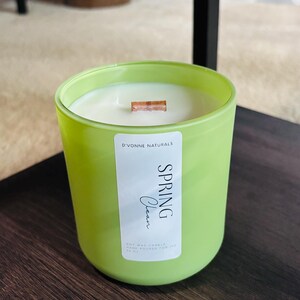 Op de afbeelding: Een groene glazen kandelaar met een witte sojawas kaars. De kaars heeft een houten lont en een label met de tekst "Spring Clean" van D'Vonne Naturals. De kaars staat op een donkerbruin oppervlak.