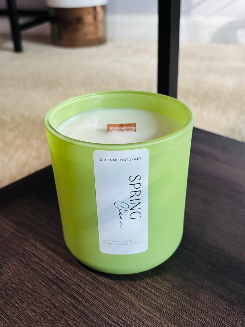 Op de afbeelding: Een groene glazen kandelaar met een witte sojawas kaars. De kaars heeft een houten lont en een label met de tekst "Spring Clean" van D'Vonne Naturals. De kaars staat op een donkerbruin oppervlak.