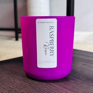 Op de afbeelding: Een helder magenta sojawas kaars in een cilindrische glazen container. Het etiket luidt "Raspberry Rose" en "D'Vonne Naturals". De kaars is met de hand gegoten en bevat 340g was. De kaars staat op een donker houten oppervlak.
