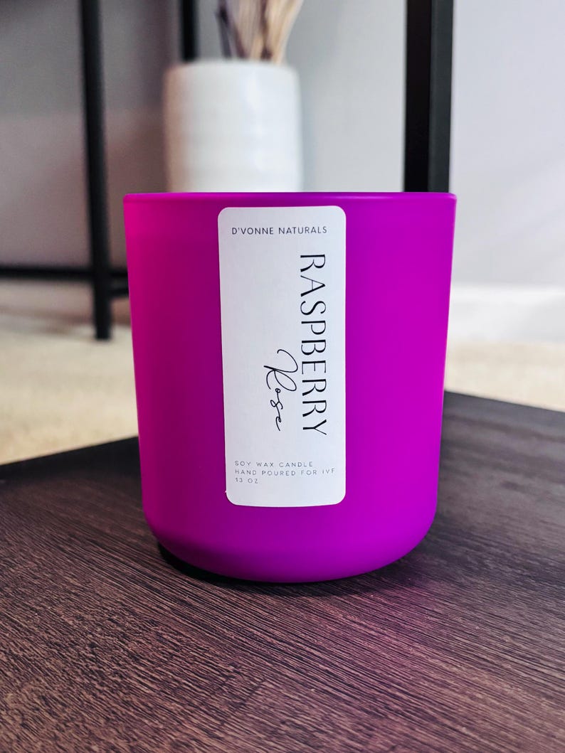 Op de afbeelding: Een helder magenta sojawas kaars in een cilindrische glazen container. Het etiket luidt "Raspberry Rose" en "D'Vonne Naturals". De kaars is met de hand gegoten en bevat 340g was. De kaars staat op een donker houten oppervlak.