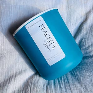 Op de afbeelding: Een turquoise sojawas kaars in een cilindrische houder met een witte rand. Het etiket luidt "Peaceful Rain" en "D'vonne Naturals". De kaars is met de hand gegoten en weegt 369 gram.