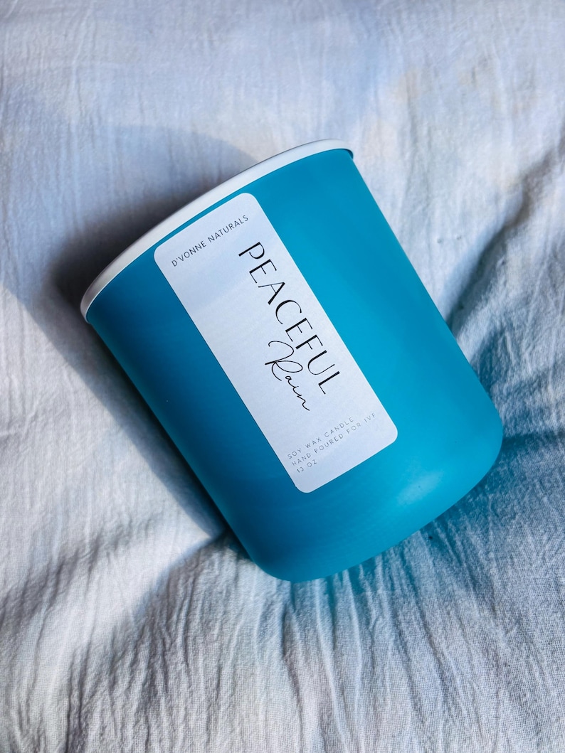 Op de afbeelding: Een turquoise sojawas kaars in een cilindrische houder met een witte rand. Het etiket luidt "Peaceful Rain" en "D'vonne Naturals". De kaars is met de hand gegoten en weegt 369 gram.