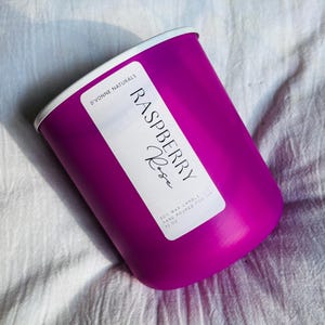 Op de afbeelding: Een magenta kaars met een witte rand, met het opschrift "Raspberry Rose". De kaars is handgegoten sojawas en weegt 312 gram. De kaars staat op een wit, getextureerd oppervlak.