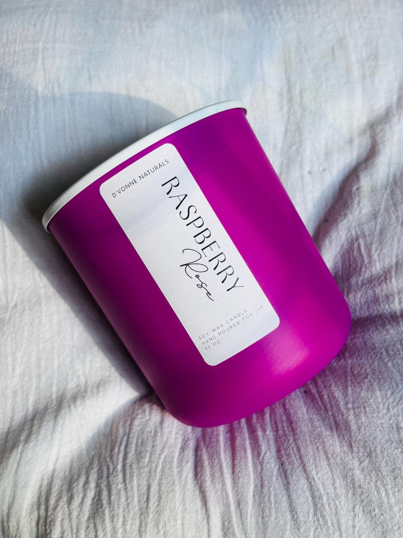 Op de afbeelding: Een magenta kaars met een witte rand, met het opschrift "Raspberry Rose". De kaars is handgegoten sojawas en weegt 312 gram. De kaars staat op een wit, getextureerd oppervlak.
