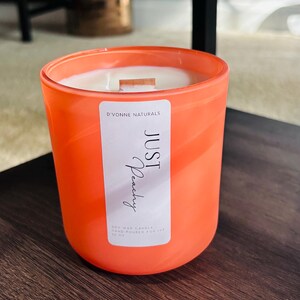 Op de afbeelding: Een oranje kaars in een glazen pot. De kaars heeft een wit etiket met de tekst "JUST Peachy" en "D'VONNE NATURALS". De kaars is een sojawas kaars, handgegoten voor 340g.