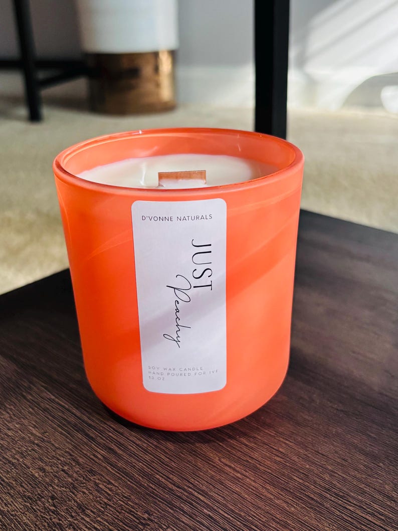 Op de afbeelding: Een oranje kaars in een glazen pot. De kaars heeft een wit etiket met de tekst "JUST Peachy" en "D'VONNE NATURALS". De kaars is een sojawas kaars, handgegoten voor 340g.
