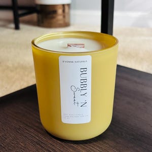 Op de afbeelding: Een gele glazen kaars met een wit etiket met de tekst "Bubbly 'N Sweet" en "D'Vonne Naturals". De kaars heeft een houten lont en is gevuld met was. De kaars is 425 gram.