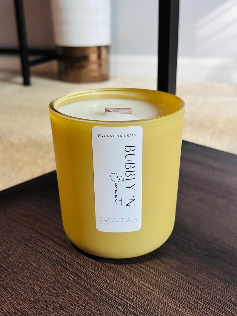 Op de afbeelding: Een gele glazen kaars met een wit etiket met de tekst "Bubbly 'N Sweet" en "D'Vonne Naturals". De kaars heeft een houten lont en is gevuld met was. De kaars is 425 gram.