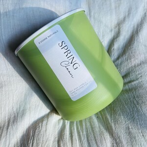 Op de afbeelding: Een groene kaars in een cilindrische houder met een witte rand. Het etiket luidt "Spring Clean". De kaars is met de hand gegoten en weegt 368,5 gram.