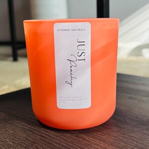Op de afbeelding: Een oranje sojawas kaars in een cilindrische glazen container. Het etiket luidt "JUST Peachy" en "D'VONNE NATURALS". De kaars is met de hand gegoten en weegt 369 gram.