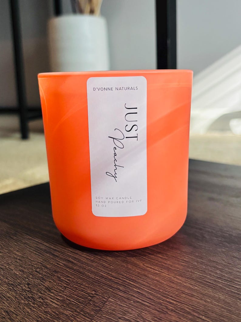 Op de afbeelding: Een oranje sojawas kaars in een cilindrische glazen container. Het etiket luidt "JUST Peachy" en "D'VONNE NATURALS". De kaars is met de hand gegoten en weegt 369 gram.