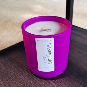 Op de afbeelding: Een helder magenta kaars in een ronde glazen container. De kaars heeft een wit wasinterieur met een houten lont. Een label op de voorkant leest "Raspberry Rose" en "D'Vonne Naturals".