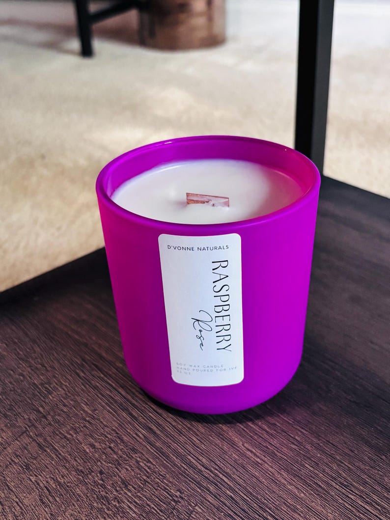 Op de afbeelding: Een helder magenta kaars in een ronde glazen container. De kaars heeft een wit wasinterieur met een houten lont. Een label op de voorkant leest "Raspberry Rose" en "D'Vonne Naturals".