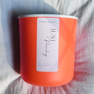 Op de afbeelding: Een feloranje sojawas kaars in een cilindrische container met een witte rand. Het etiket luidt "JUST Peachy" en "D'VONNE NATURALS". De kaars is met de hand gegoten en weegt 312 gram.