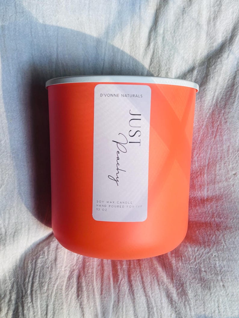Op de afbeelding: Een feloranje sojawas kaars in een cilindrische container met een witte rand. Het etiket luidt "JUST Peachy" en "D'VONNE NATURALS". De kaars is met de hand gegoten en weegt 312 gram.
