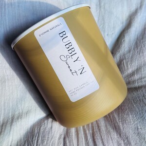 Op de afbeelding: Een goudkleurige cilindrische kaars met een witte rand. Het etiket luidt "BUBBLY 'N Sweet" en "SOY WAX CANDLE HAND POURED FOR IVF 13 OZ" in zwarte tekst. De kaars staat tegen een witte stoffen achtergrond.