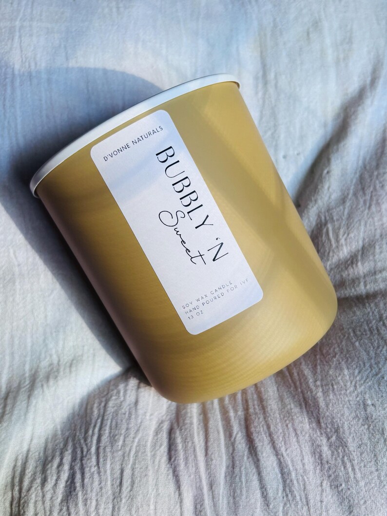 Op de afbeelding: Een goudkleurige cilindrische kaars met een witte rand. Het etiket luidt "BUBBLY 'N Sweet" en "SOY WAX CANDLE HAND POURED FOR IVF 13 OZ" in zwarte tekst. De kaars staat tegen een witte stoffen achtergrond.