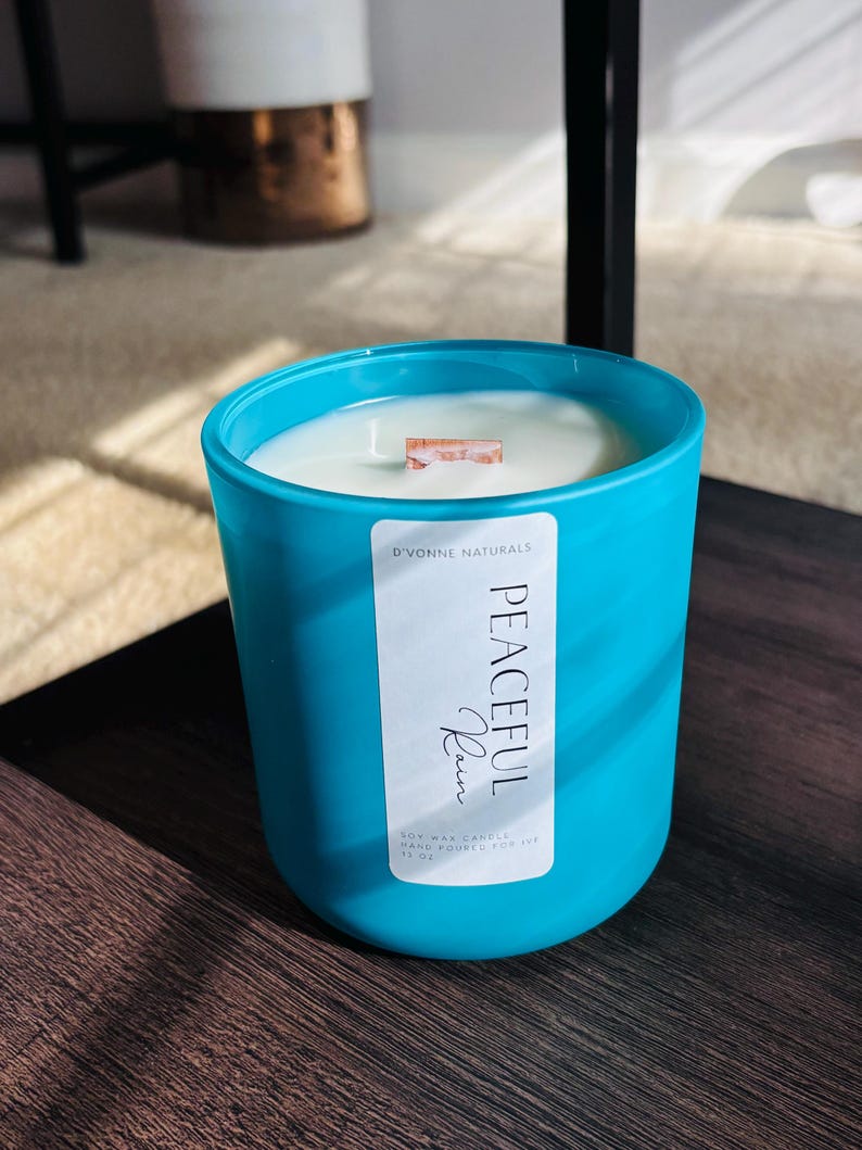 Op de afbeelding: Een turquoise sojawas kaars in een glazen pot met een houten lont. Het etiket leest "Peaceful Rain" en "D'Vonne Naturals". De kaars is met de hand gegoten en staat op een donker houten oppervlak.