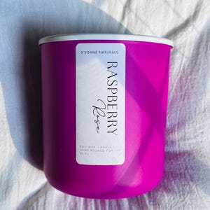 Op de afbeelding: Een helder magenta sojawas kaars in een cilindrische container met een witte rand. Het label luidt "Raspberry Rose" en "D'Vonne Naturals". De kaars is met de hand gegoten en weegt 369 gram.