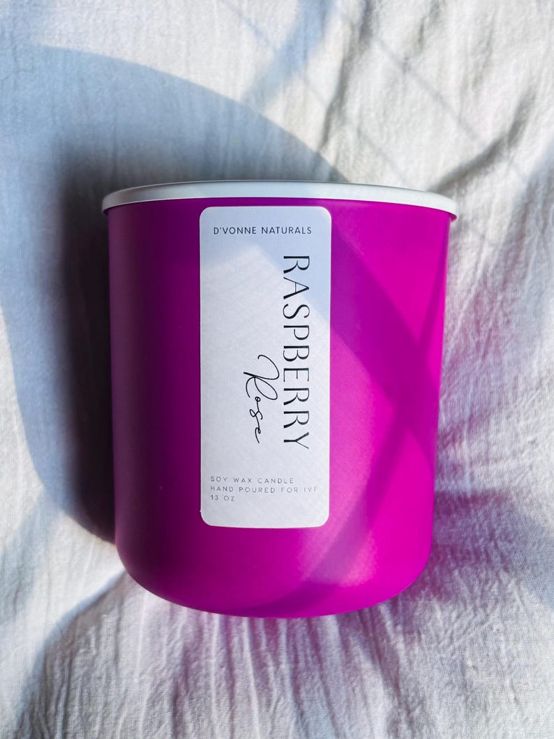 Op de afbeelding: Een helder magenta sojawas kaars in een cilindrische container met een witte rand. Het label luidt "Raspberry Rose" en "D'Vonne Naturals". De kaars is met de hand gegoten en weegt 369 gram.