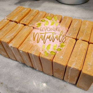 May include: Orange-colored rectangular bars of soap, individually wrapped in clear plastic. The soap bars are arranged in neat rows. The image includes the text "D'VONNE NATURALS" in gold lettering with a pink background and green leaves.