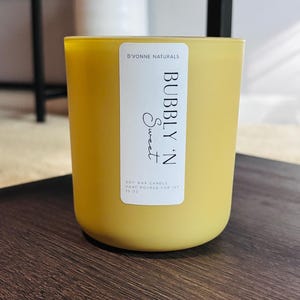 Op de afbeelding: Een gele sojawas kaars in een glazen pot. Het etiket luidt "D'VONNE NATURALS BUBBLY 'N Sweet" en "HAND POURED FOR IVF 369g". De kaars heeft een afgeronde vorm en staat op een donker houten oppervlak.