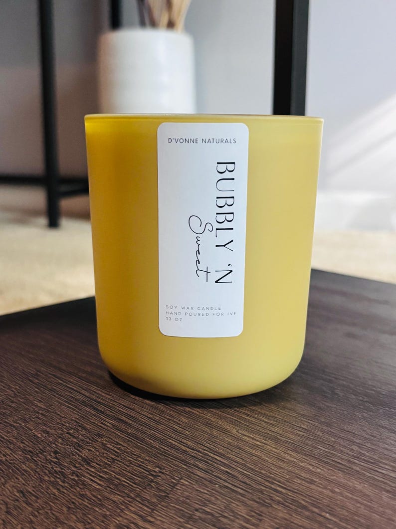 Op de afbeelding: Een gele sojawas kaars in een glazen pot. Het etiket luidt "D'VONNE NATURALS BUBBLY 'N Sweet" en "HAND POURED FOR IVF 369g". De kaars heeft een afgeronde vorm en staat op een donker houten oppervlak.
