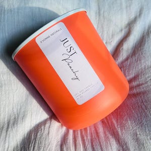 Op de afbeelding: Een oranje kaars met een witte rand en een wit etiket met de tekst "JUST Peachy" in script. Het etiket bevat ook de tekst "D'VONNE NATURALS" en "SOY WAX CANDLE HAND POURED FOR 13 OZ". De kaars staat op een wit, getextureerd oppervlak.
