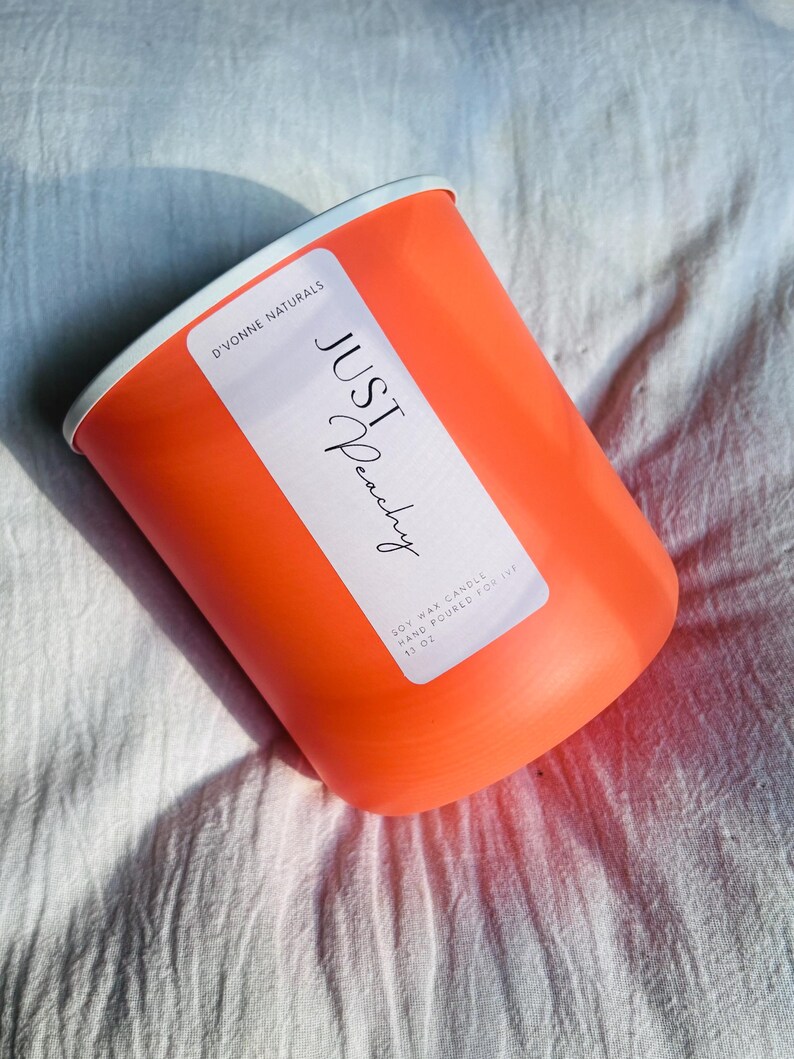 Op de afbeelding: Een oranje kaars met een witte rand en een wit etiket met de tekst "JUST Peachy" in script. Het etiket bevat ook de tekst "D'VONNE NATURALS" en "SOY WAX CANDLE HAND POURED FOR 13 OZ". De kaars staat op een wit, getextureerd oppervlak.
