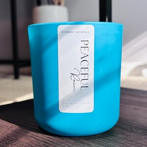 Op de afbeelding: Een turquoise sojawas kaars in een glazen pot. Het etiket luidt "PEACEFUL Rain" en "D'VONNE NATURALS". De kaars is met de hand gegoten en bevat 369 gram was. De kaars staat op een donker houten oppervlak.