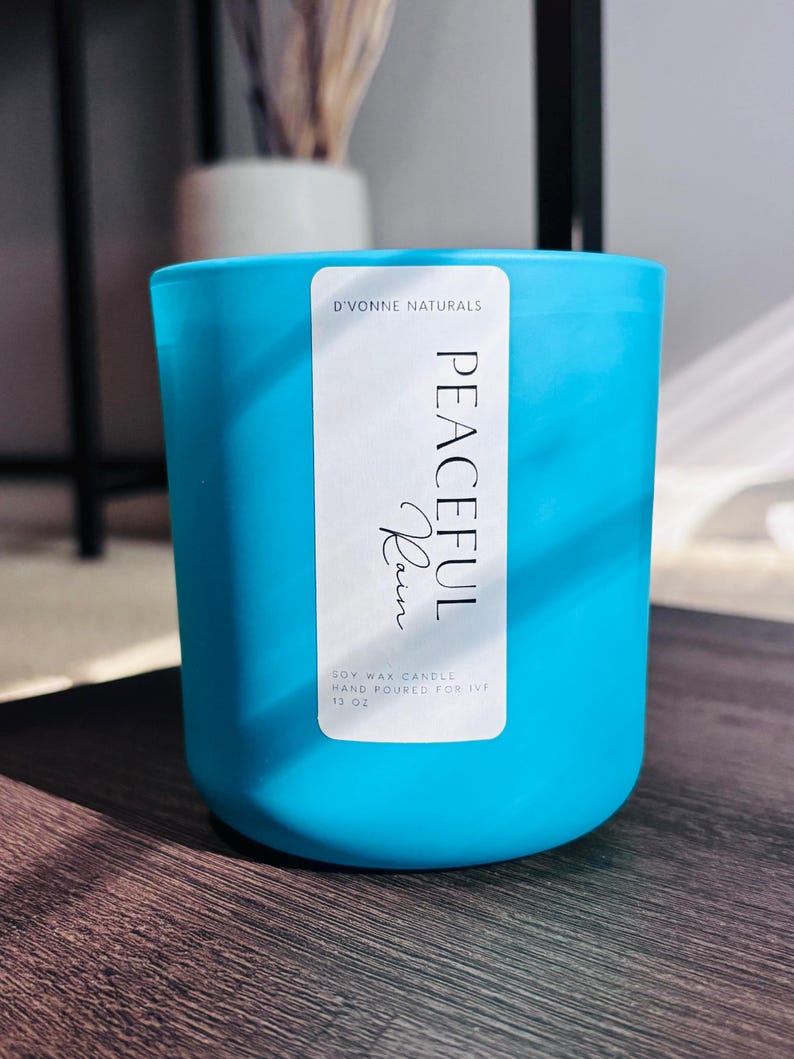 Op de afbeelding: Een turquoise sojawas kaars in een glazen pot. Het etiket luidt "PEACEFUL Rain" en "D'VONNE NATURALS". De kaars is met de hand gegoten en bevat 369 gram was. De kaars staat op een donker houten oppervlak.