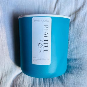 Op de afbeelding: Een turquoise sojawas kaars in een cilindrische houder met een witte rand. Het etiket leest "PEACEFUL Rain" en "D'VONNE NATURALS". De kaars is met de hand gegoten en weegt 369 gram.