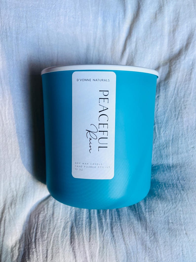 Op de afbeelding: Een turquoise sojawas kaars in een cilindrische houder met een witte rand. Het etiket leest "PEACEFUL Rain" en "D'VONNE NATURALS". De kaars is met de hand gegoten en weegt 369 gram.
