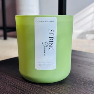 Op de afbeelding: Een groene sojawas kaars in een glazen pot. Het etiket luidt "D'Vonne Naturals SPRING Clean" en "Hand Poured for IVP 13 oz". De kaars staat op een donker houten oppervlak. Een witte vaas staat op de achtergrond.