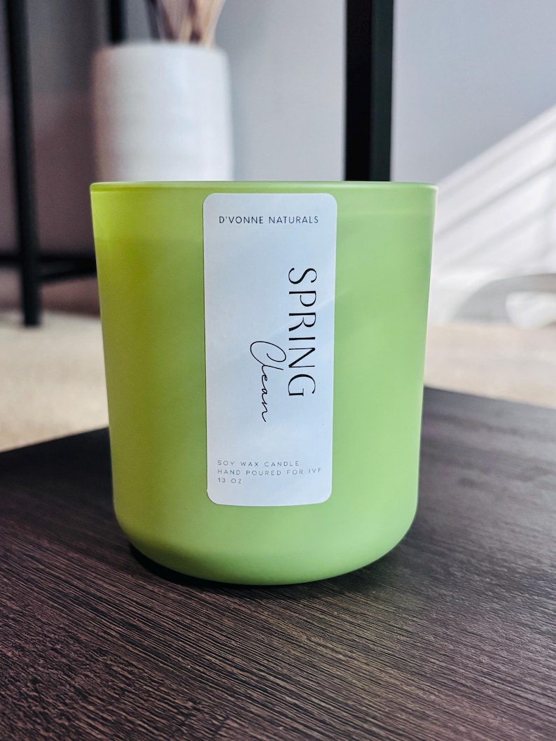 Op de afbeelding: Een groene sojawas kaars in een glazen pot. Het etiket luidt "D'Vonne Naturals SPRING Clean" en "Hand Poured for IVP 13 oz". De kaars staat op een donker houten oppervlak. Een witte vaas staat op de achtergrond.