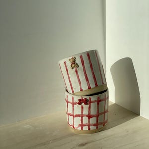 Könnte beinhalten: Zwei weiße Keramikbecher mit roten und weißen Mustern. Der obere Becher hat rote Streifen und eine kleine braune Bärenfigur. Der untere Becher hat ein rotes und weißes Karomuster mit einer roten Schleife.