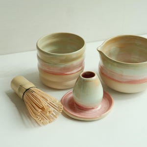 Könnte beinhalten: Ein Matcha-Teeset aus Keramik mit einer Schale mit Ausguss, einem zylindrischen Behälter und einem kleinen Besenhalter auf einer Untertasse, alles in einem Farbverlauf aus Creme, Blassgrün und Altrosa gehalten, inklusive eines Bambusbesens.